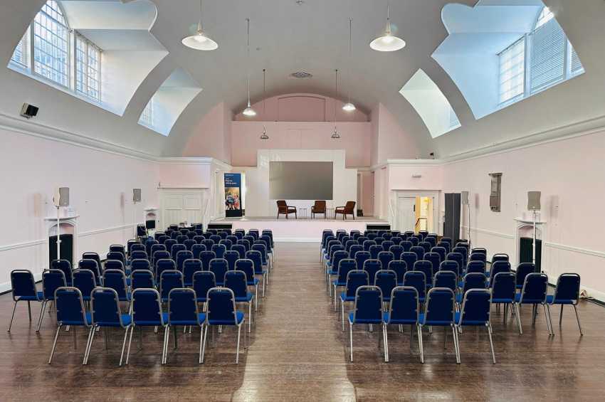 Mary Ward Hall set up with rows of blue chairs facing a stage, skylights overhead, and pendant lighting.