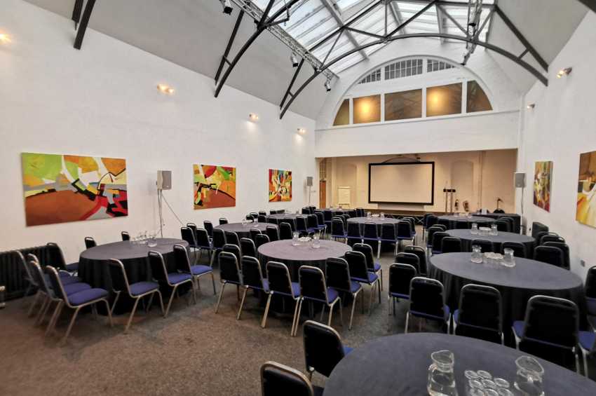 Mary Ward Hall set up with round tables, black chairs, blue seat pads, and projection screen under vaulted glass ceiling.