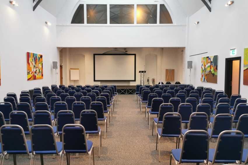 Large hall with rows of blue chairs facing a projection screen and stage area, modern lighting and artwork on walls.