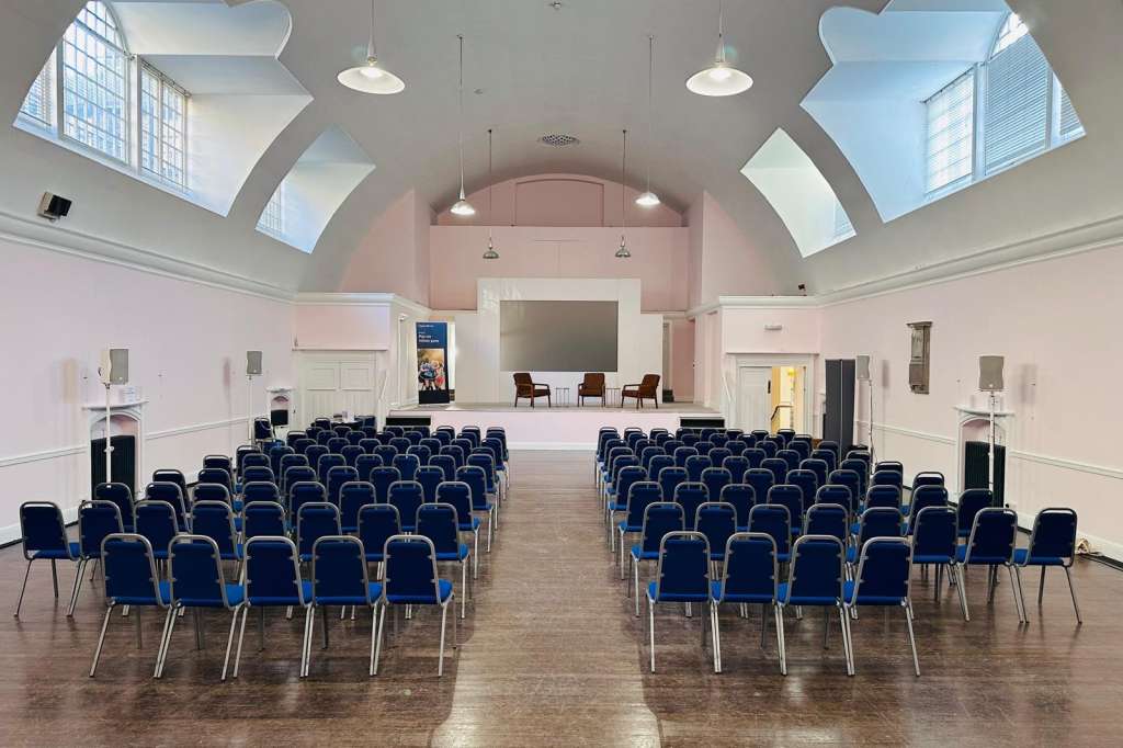 Mary Ward Hall set up with rows of blue chairs facing a stage, skylights overhead, and pendant lighting.