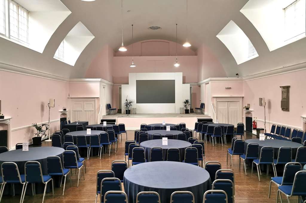 Mary Ward Hall set up with round tables, black chairs, and a stage with projection screen and pendant lighting.