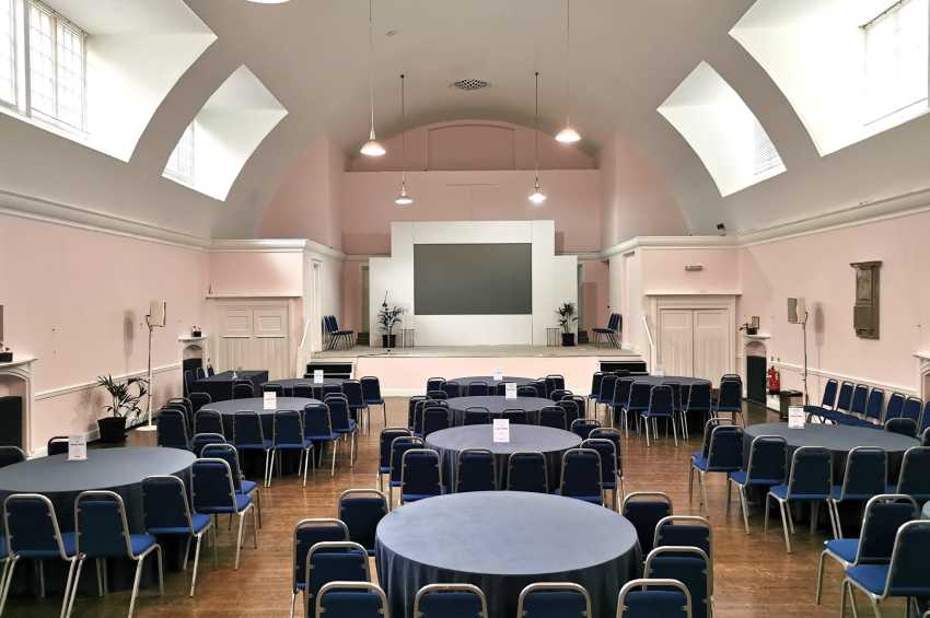 Mary Ward Hall set up with round tables, black chairs, and a stage with projection screen and pendant lighting.
