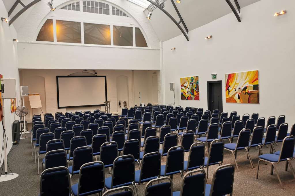 Mary Ward Hall set up with rows of blue-cushioned chairs facing a projection screen and stage area.