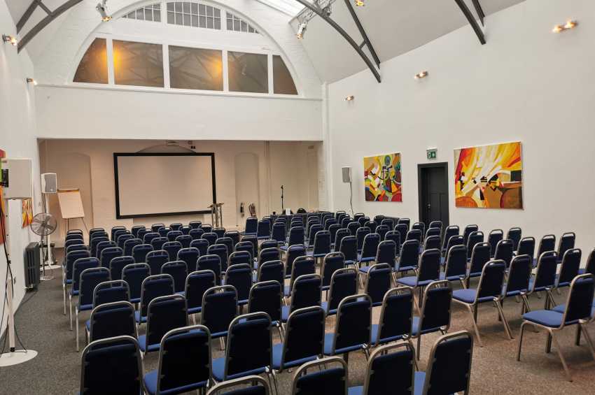 Mary Ward Hall set up with rows of blue-cushioned chairs facing a projection screen and stage area.