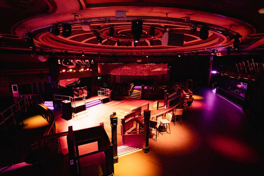 Modern event space with circular neon ceiling, stage, scattered seating, and dramatic red and purple lighting.