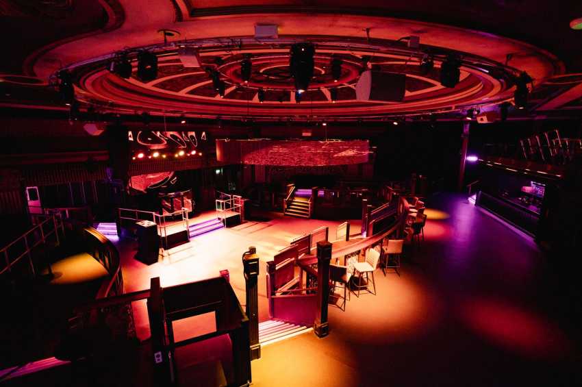 Modern event space with circular neon ceiling, stage, scattered seating, and dramatic red and purple lighting.