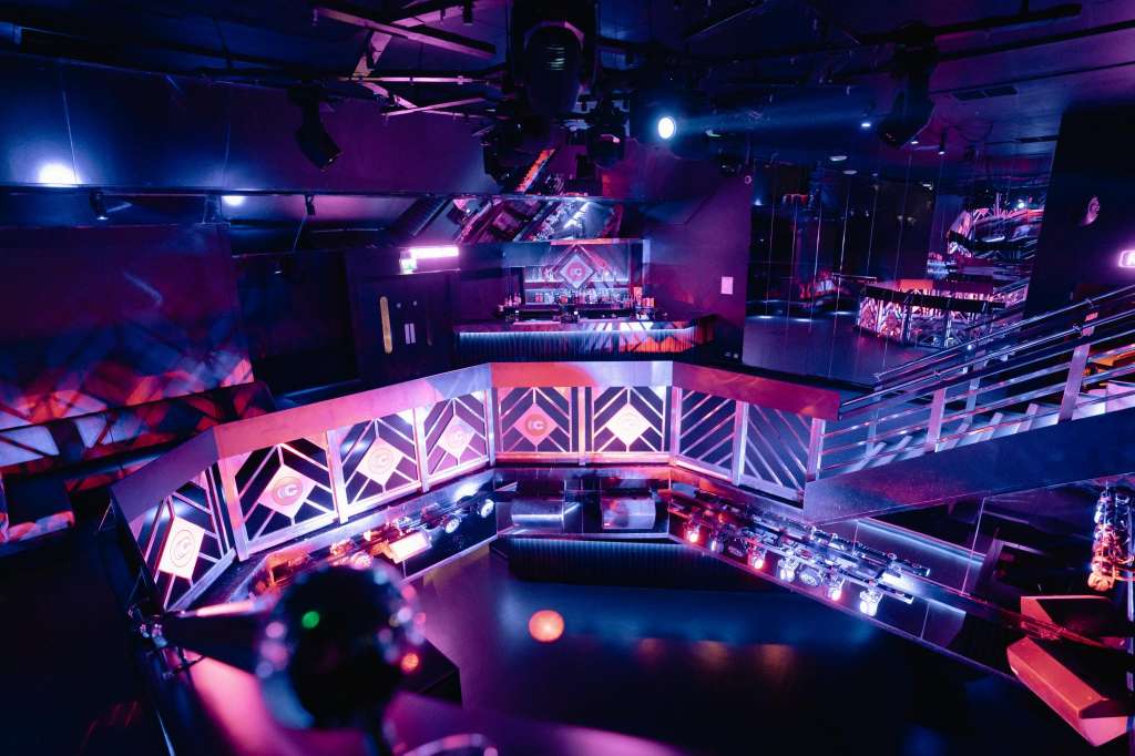 Modern event space with elevated stage, geometric LED panels, blue and pink lighting, and tiered seating areas.