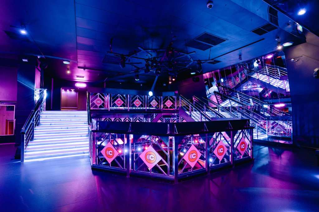 Modern event space with blue and purple lighting, illuminated staircase, multi-level gallery, and branded display screens.