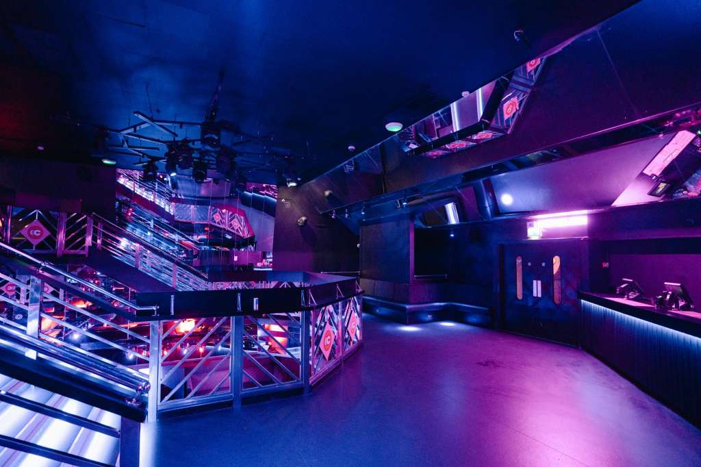Modern event space with multi-level platform, neon pink and blue lighting, industrial ceiling rig, and bar counter.
