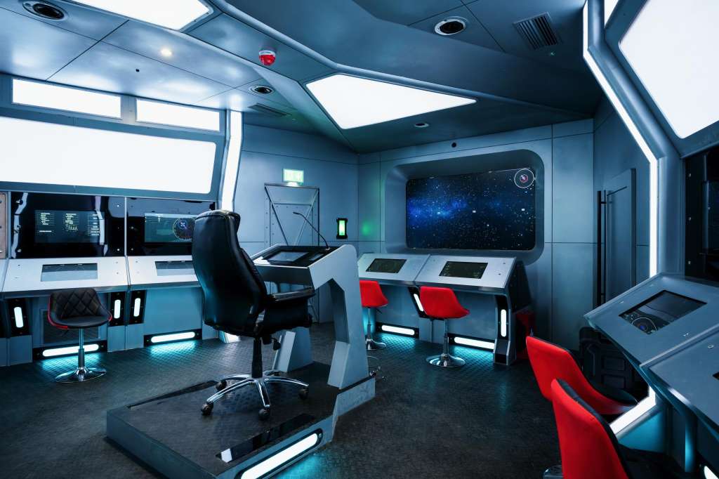 Futuristic control room with blue lighting, multiple workstations, red chairs, and starfield display screen.