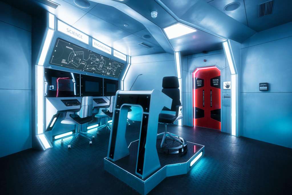 Futuristic interior with neon-lit gaming stations, control panels, and red emergency door under blue lighting.