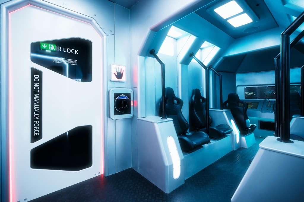 Futuristic escape room with neon-lit air lock door, gaming seats, and blue ambient lighting throughout.