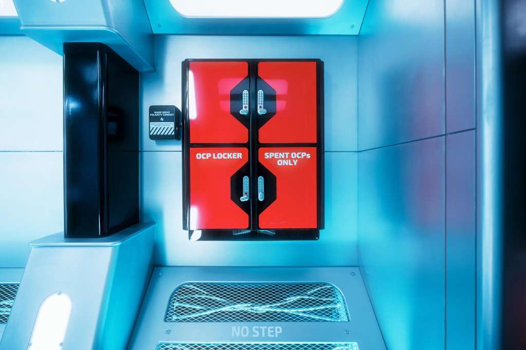Interior of a shipping container with blue walls, red locker doors labeled OCP LOCKER and SPENT OCPs ONLY, and industrial equipment.