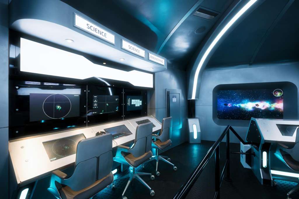 High-tech control room with multiple illuminated displays, workstations, and blue ambient lighting throughout.