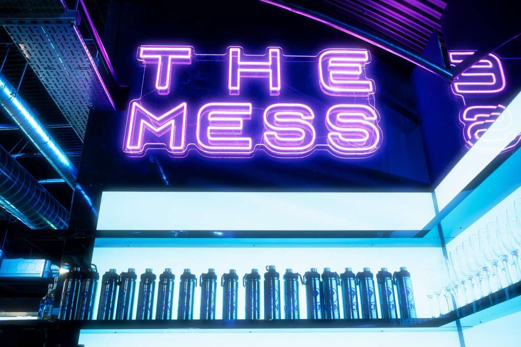 Neon sign reading 'THE MESS' above illuminated shelving with bottles in blue and purple lighting.