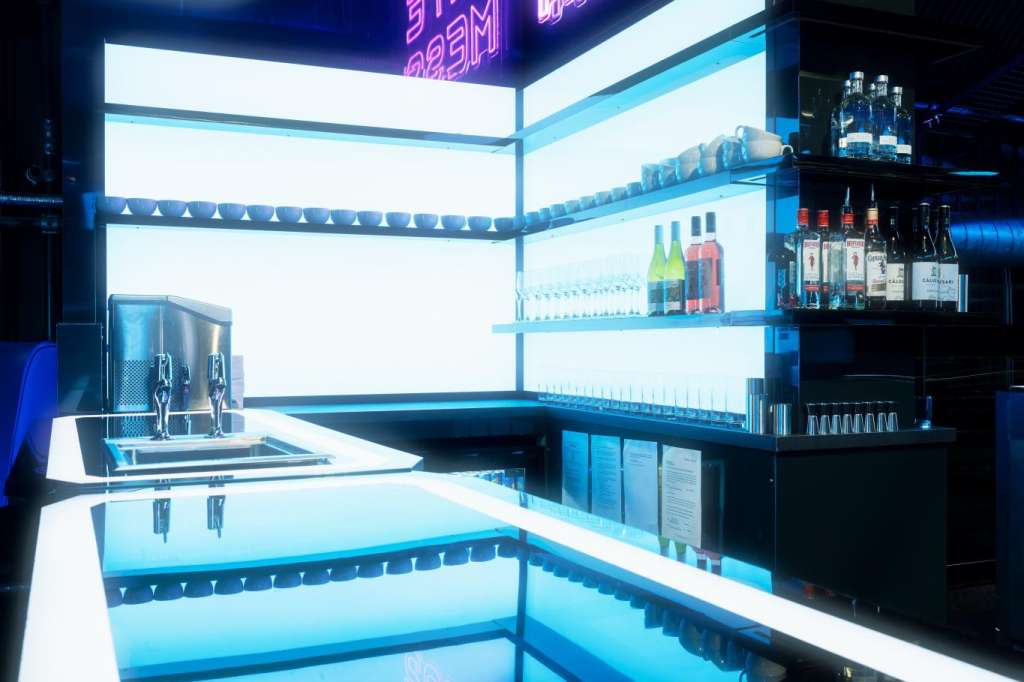 Modern bar counter with neon signage, illuminated shelving displaying bottles, and blue ambient lighting throughout.