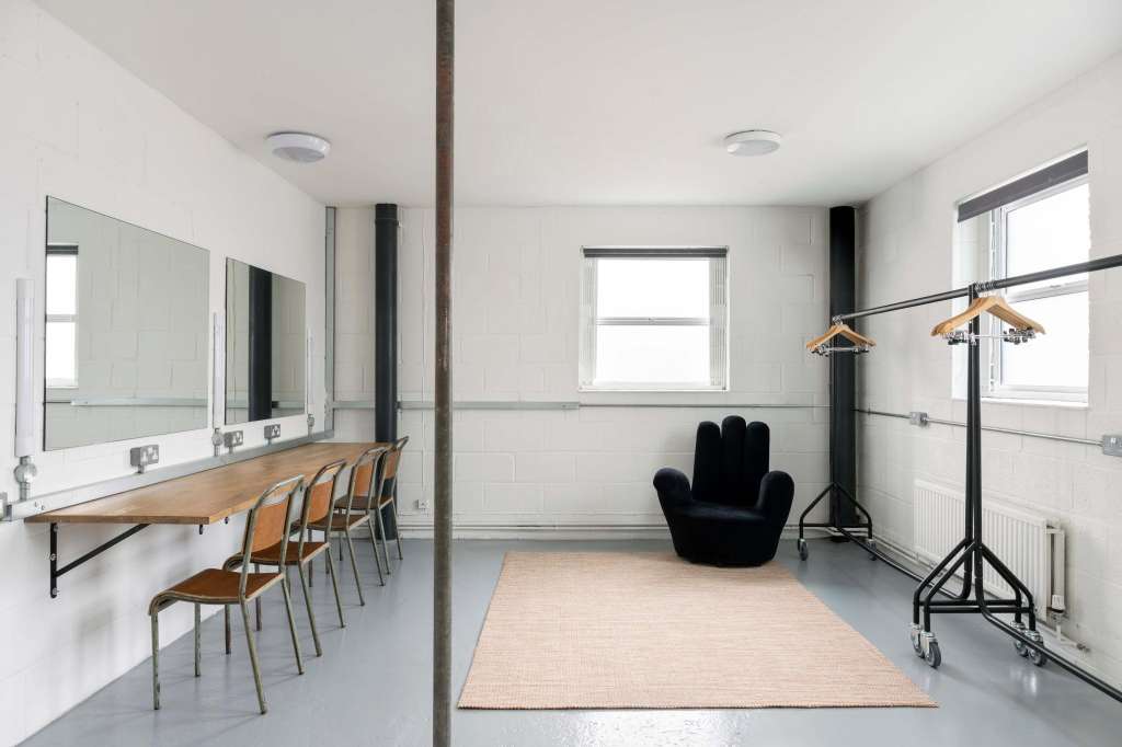 Modern studio space with makeup vanity, mirrors, wooden chairs, black armchair, and professional lighting equipment on tripods.