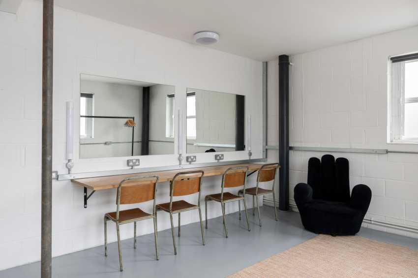 Modern studio space with vanity mirrors, wooden chairs, black sculptural seating, and polished concrete flooring.