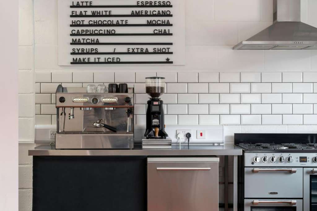 Café kitchen counter with espresso machine, menu board, white subway tile backsplash, and orange-black checkered flooring.