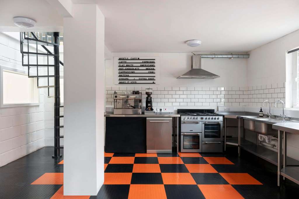 Commercial kitchen with stainless steel appliances, white subway tile backsplash, menu board, and orange-black checkered flooring.