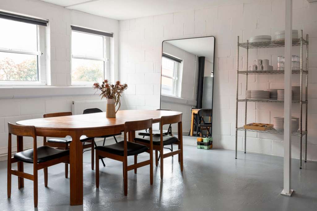 Bright studio space with wooden dining table, mid-century chairs, large windows, dried flowers, and metal shelving.