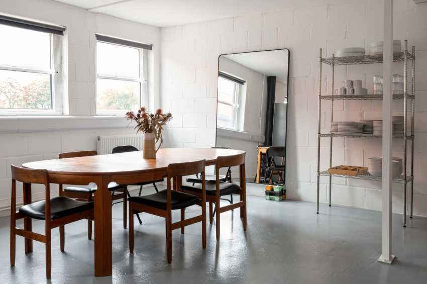 Bright studio space with wooden dining table, mid-century chairs, large windows, dried flowers, and metal shelving.