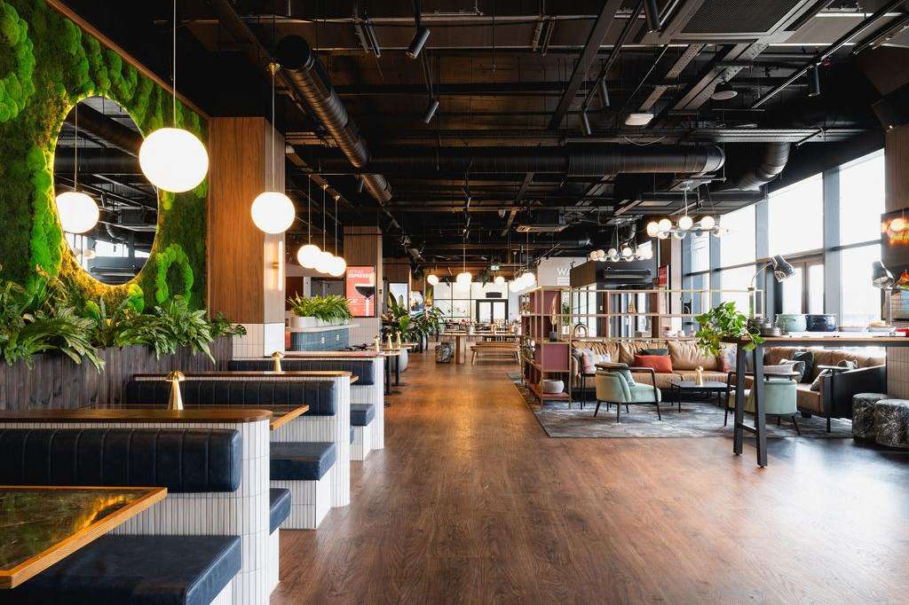 Modern gallery space with exposed ceiling, pendant lighting, green living wall, wood accents, and lounge seating areas.