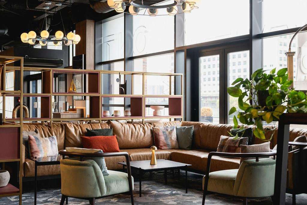 The Gallery lounge area with brown leather sectional, green armchairs, modern pendant lighting, and floor-to-ceiling windows overlooking city.