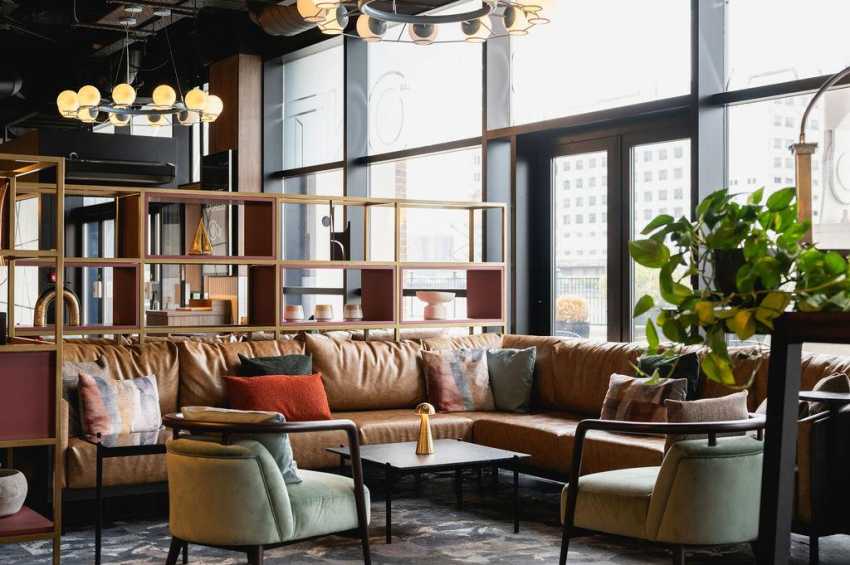 The Gallery lounge area with brown leather sectional, green armchairs, modern pendant lighting, and floor-to-ceiling windows overlooking city.