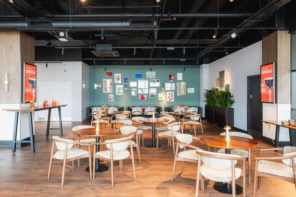 Gallery space with round wooden tables, mid-century chairs, teal accent wall with artwork, and modern lighting.