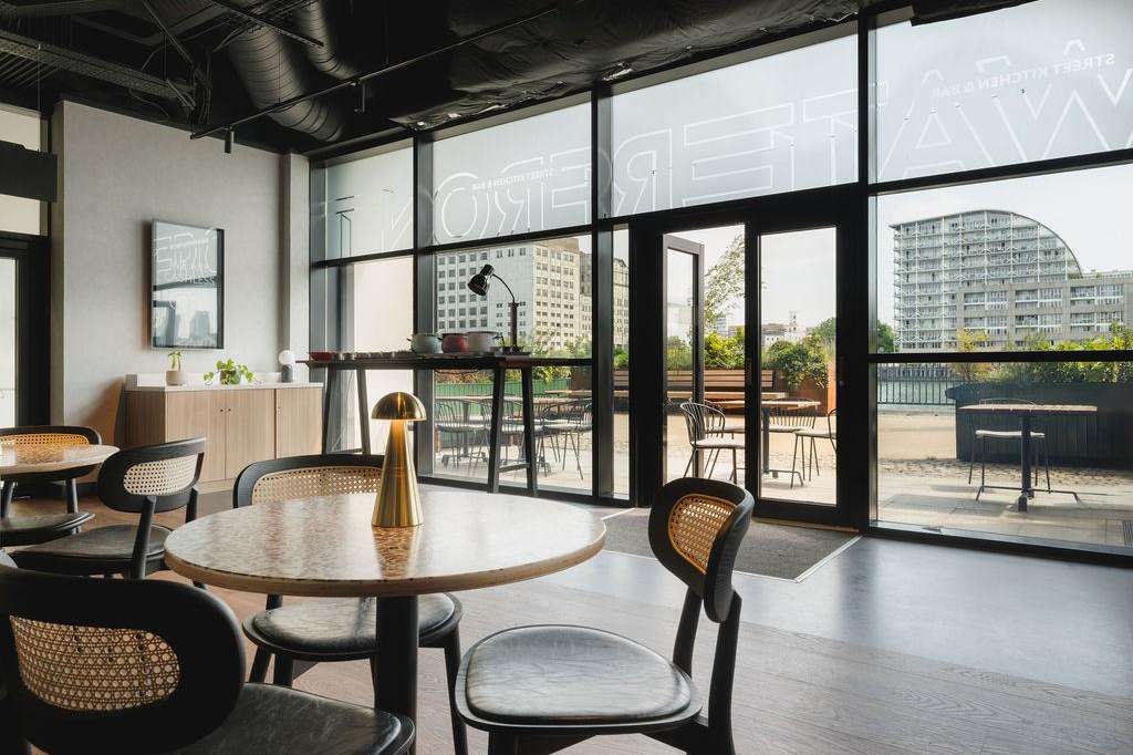 Modern gallery space with round tables, contemporary chairs, floor-to-ceiling windows overlooking a rooftop terrace and city skyline.