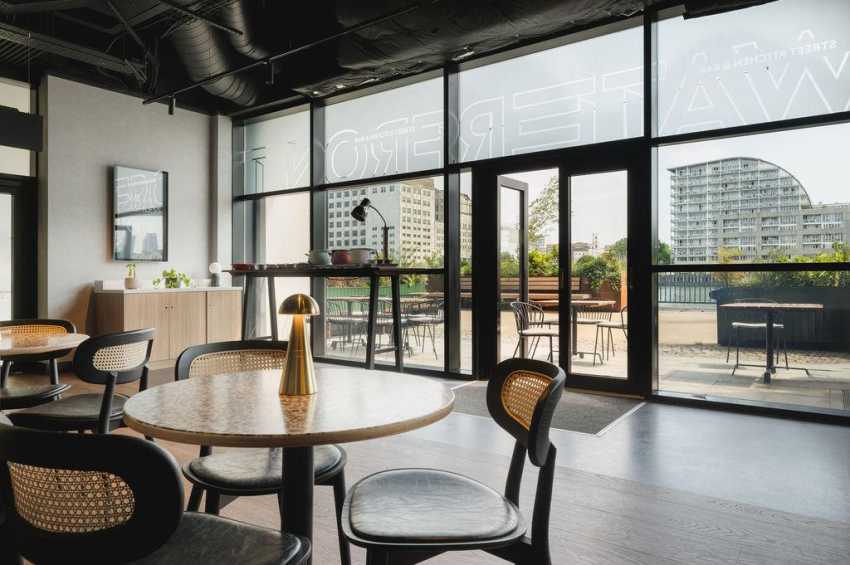 Modern gallery space with round tables, contemporary chairs, floor-to-ceiling windows overlooking a rooftop terrace and city skyline.
