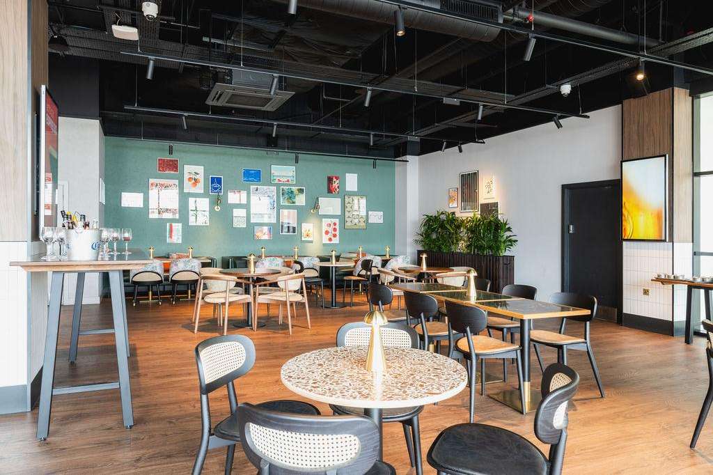 Modern gallery space with scattered tables, mixed seating, bar counter, and colorful artwork on turquoise and white walls.