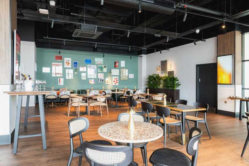 Modern gallery space with scattered tables, mixed seating, bar counter, and colorful artwork on turquoise and white walls.