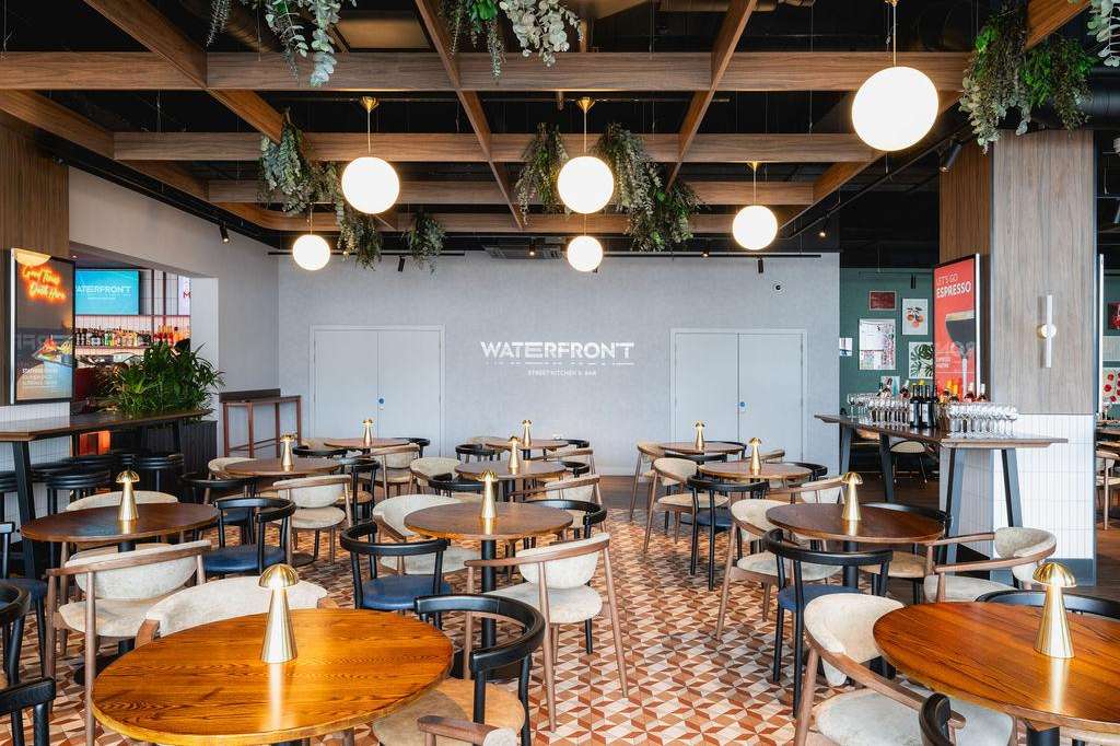 Modern dining space with round wooden tables, cream chairs, hanging greenery, globe pendant lights, and geometric floor pattern.