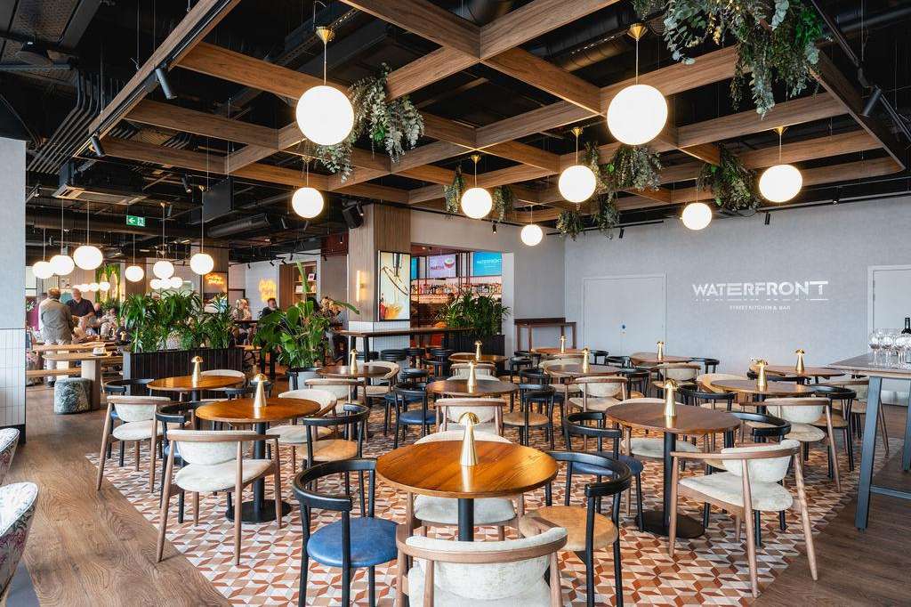 Modern dining space with round wooden tables, mid-century chairs, hanging globe lights, and cascading greenery from wooden ceiling beams.