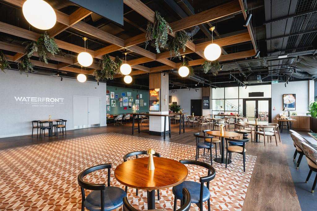 Modern gallery space with round tables, wooden beams, pendant lighting, and hanging greenery. Exposed industrial ceiling.