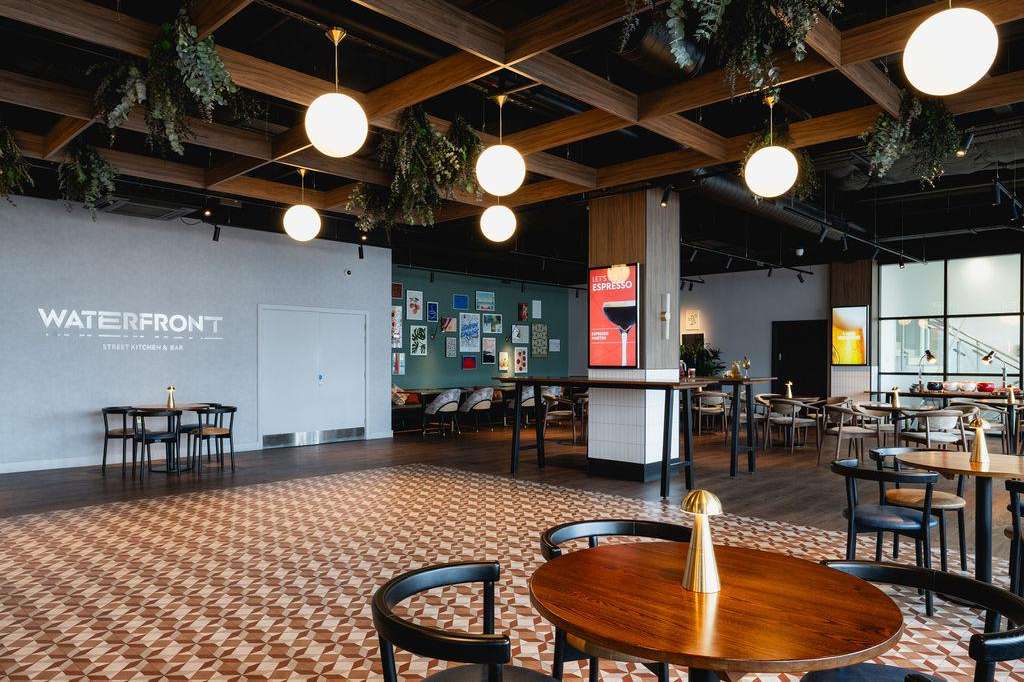 Modern gallery space with wooden ceiling beams, pendant lighting, scattered tables, and green foliage accents.