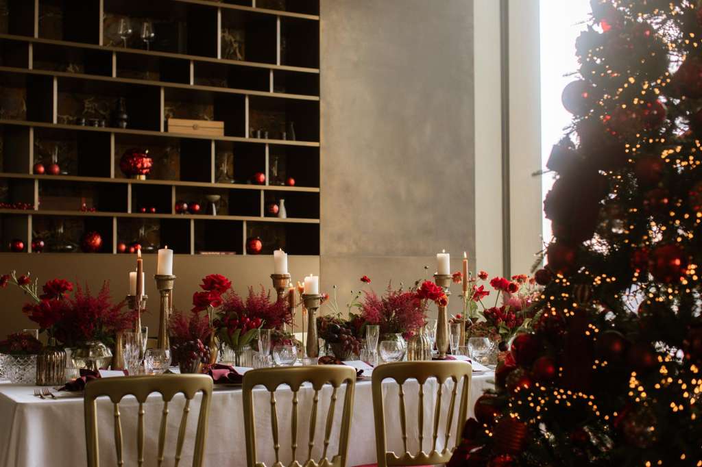 Ballroom set for a formal dinner with round tables, red floral centerpieces, candlesticks, and a decorated Christmas tree.