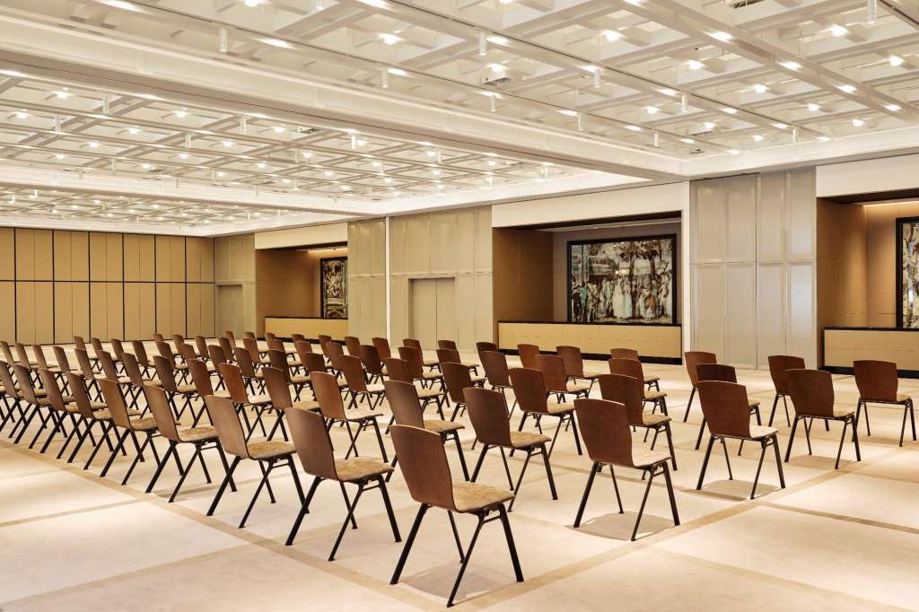 Meeting room with rows of brown chairs facing a stage, coffered ceiling with recessed lighting, and neutral wood paneling.