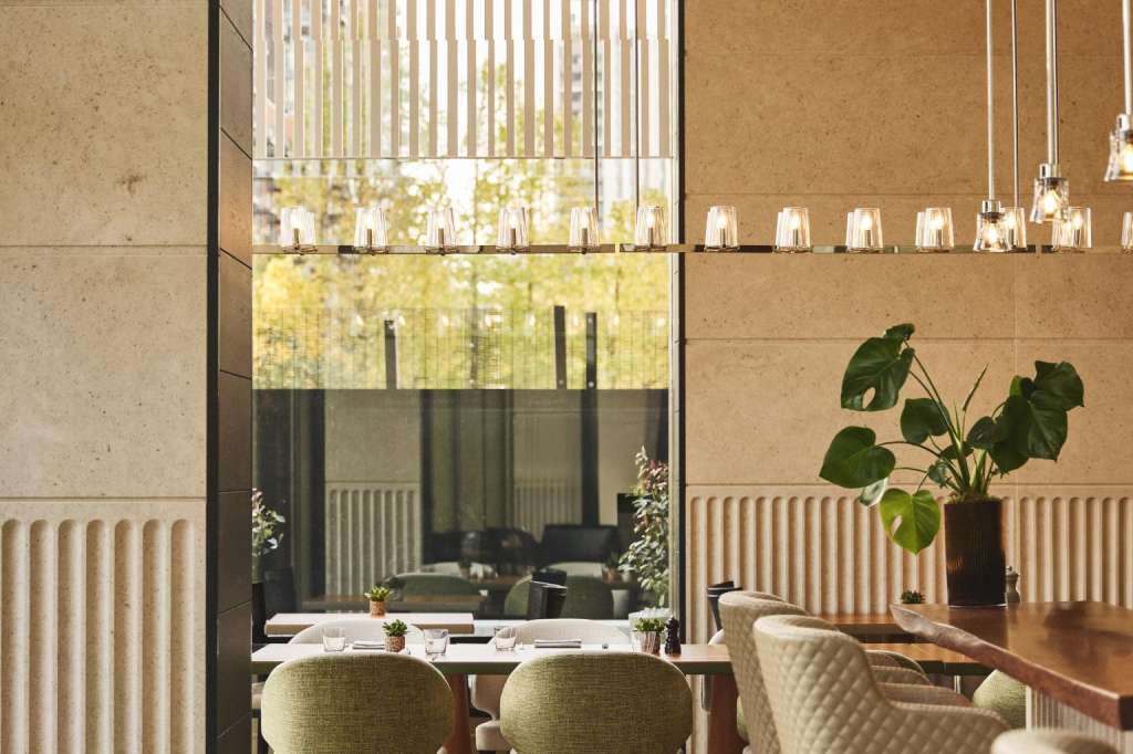 Modern dining space with floor-to-ceiling windows, pendant lighting, green upholstered chairs, and potted plants.