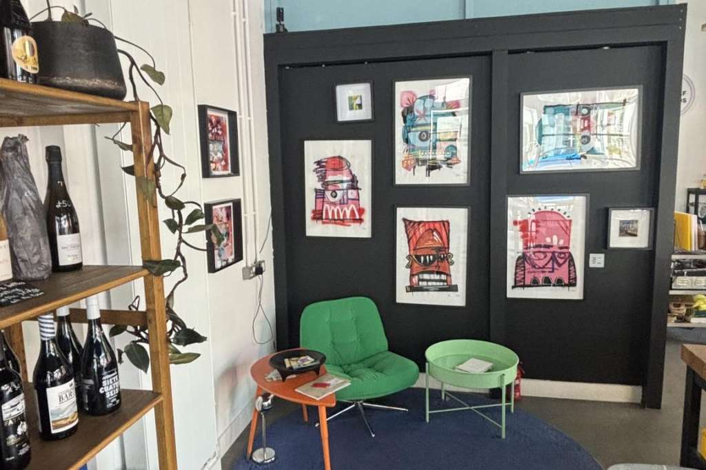 Interior space with art display wall, green chairs, round blue rug, wooden shelving with bottles and merchandise.