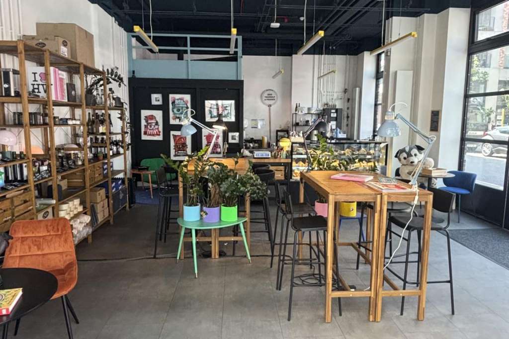 Industrial loft space with high ceilings, exposed ductwork, wooden bar tables, colorful chairs, bookshelves, and natural light from large windows.