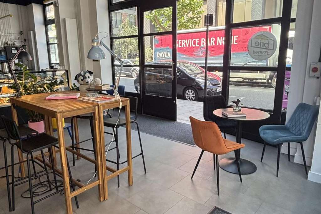 Modern café interior with high ceilings, wooden bar table, black bistro tables, and large storefront windows.