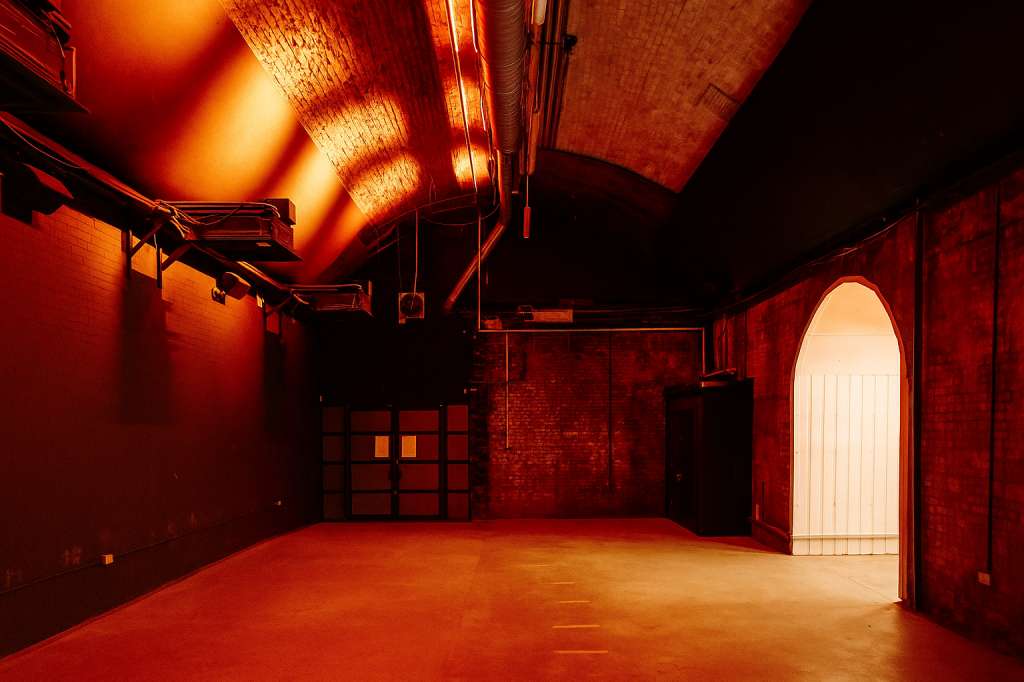 Industrial warehouse space with red brick walls, dramatic red and orange lighting, and exposed ceiling fixtures.