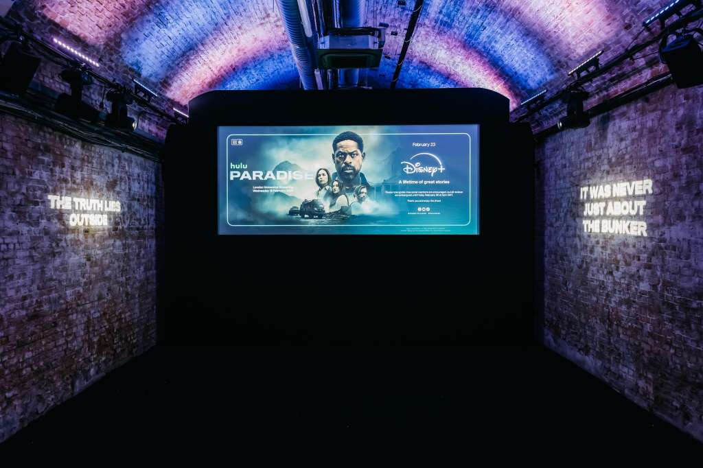 Industrial brick tunnel with atmospheric blue and purple lighting, large digital screen displaying media content.