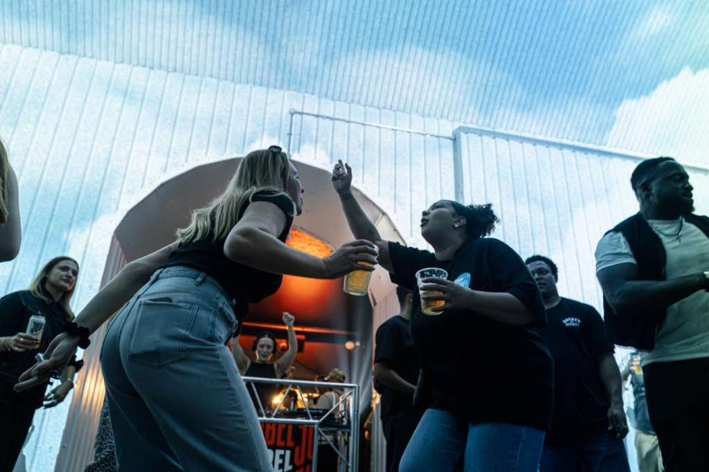 Outdoor rooftop space with industrial corrugated ceiling, groups of people holding drinks gathered around a central fire feature.