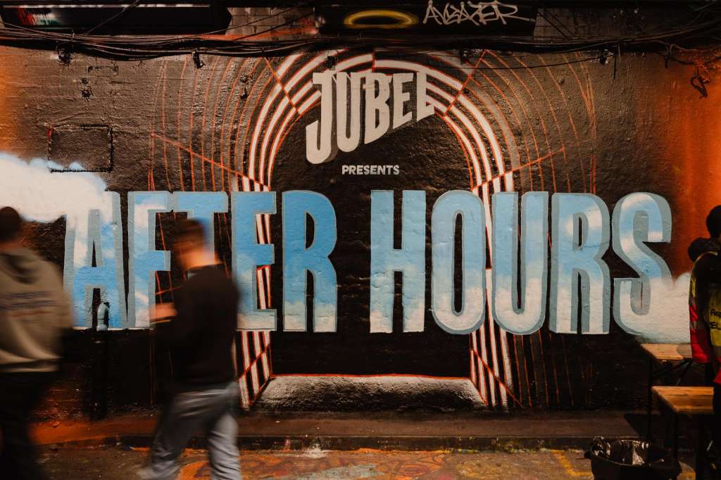 Industrial venue space with neon signage reading 'After Hours' and branded backdrop, people visible in motion.