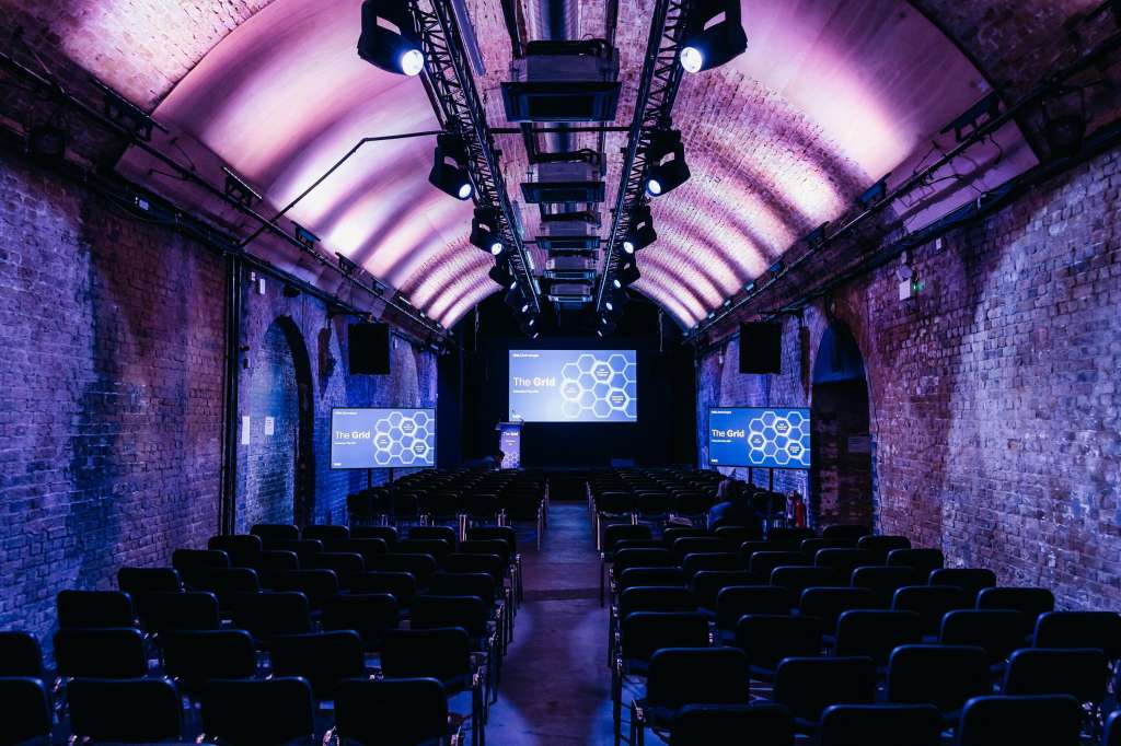 Industrial brick venue with arched ceiling, purple ambient lighting, rows of black seating, and projection screens displaying branding.