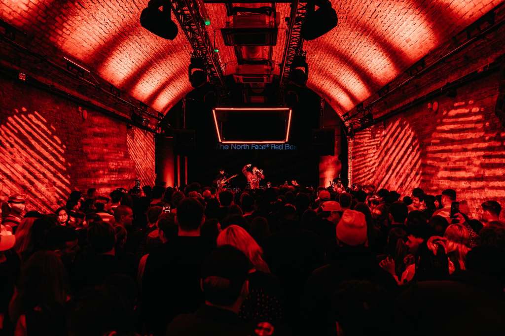 Industrial brick venue with red atmospheric lighting, crowd gathered for live performance on stage.